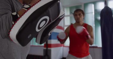 Front view of a mixed race male kickboxer practicing a kick against a pad held by a bald Caucasian male trainer in a boxing gym, slow motion