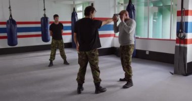 Krav Maga Training. Front view of a bald Caucasian male instructor giving self defense training in a boxing gym demonstrating a gun defense to a mixed race man, while another man looks on, slow motion