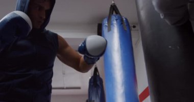 Low angle view of a mixed race male boxer punching a punchbag hold by a bald Caucasian male trainer in a boxing gym, slow motion