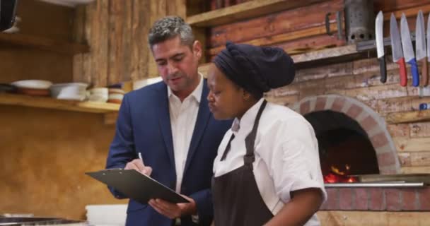 キッチンの白人男性レストランマネージャーの正面図 クリップボードを保持し アフリカ系アメリカ人女性料理人と話をし 指示を与える — ストック動画