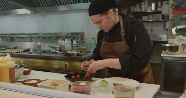 一个白人女厨师在一个繁忙的餐厅厨房里工作 把西红柿放在盘子里 她的女同事把她的平底锅和食物放在一起 在镜头前她看到了这个白人女厨师 — 图库视频影像