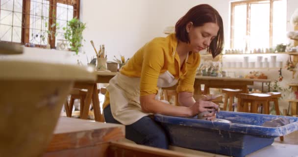 一个年轻的白种人女陶工 头戴围裙 头戴黄褐色头发 坐在陶瓷轮旁 在陶瓷工作室里翻动一块粘土 — 图库视频影像