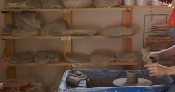 一个年轻的白种人女陶工头戴围裙 坐在陶瓷轮旁 转动锅子 在陶瓷工作室里塑造出自己的形象 她一头乌黑的头发 一头蓬蓬蓬蓬的发型 — 图库视频影像