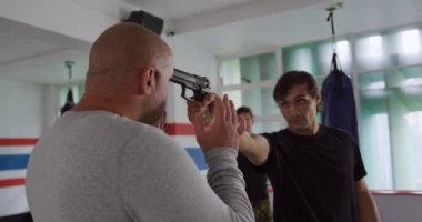 Krav Maga Training. Rear view close up of a bald Caucasian male instructor giving self defence training in a boxing gym demonstrating a gun defense to a Caucasian man, while another man looks on
