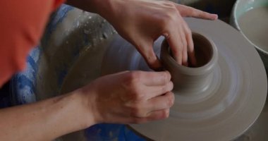 Çömlekçinin tekerinde oturan ve çömlek atölyesinde ağır çekim şekillendiren genç beyaz kadın çömlekçinin yüksek açılı görüntüsü.