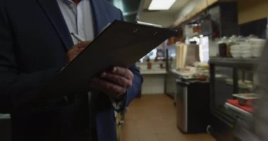 Front view mid section of a Caucasian male manager in a busy restaurant kitchen, holding a clipboard and writing, cooks working in the background