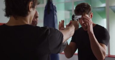 Krav Maga Eğitimi. Kafkasyalı bir adam ve karışık ırktan bir adamın, boks salonunda kel beyaz bir erkek eğitmenle silah savunması yapmasını yakından izliyoruz.