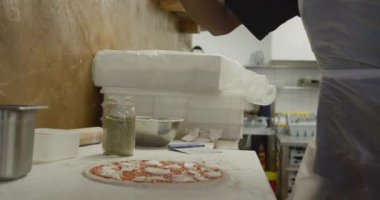 Side view mid section of a mixed race male cook working in a busy pizza restaurant kitchen, sprinkling herbs over pizza dough before cooking