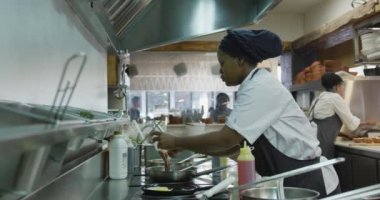 Meşgul bir restoran mutfağında çalışan, kızartma tavasında yemek yapan, arka planda çalışan Afrikalı Amerikalı bir kadın aşçının yan görüntüsü.