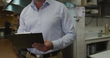 Portrait of a happy Caucasian male manager in a busy restaurant kitchen, smiling to camera holding a clipboard and writing, cooks working in the background 