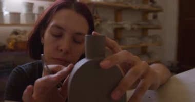 Kafkasyalı, kısa saçlı, mavi gözlü genç bir çömlekçinin ön görüntüsü. Bir tencere tutuyor ve arka planda rafları olan bir çömlek atölyesinde parmaklarıyla çalışıyor.