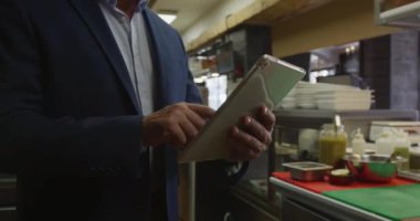 Front view mid section of a Caucasian male manager in a busy restaurant kitchen, holding a tablet computer, cooks working in the background