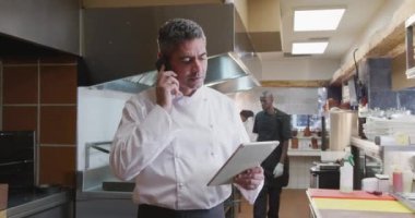 Meşgul bir lokanta mutfağında çalışan, elinde tablet bilgisayar olan ve arka planda çalışan, akıllı telefondan konuşan beyaz bir aşçının ön görüntüsü.