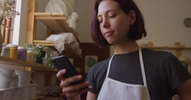 一位白种人年轻的女诗人 一头乌黑的头发 发型蓬松 站在窗边的陶瓷工作室里 用智能手机向窗外望去 动作缓慢 — 图库视频影像