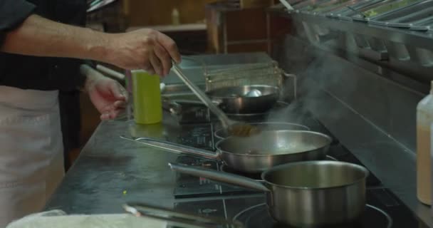 忙しいレストランのキッチンで働く白人男性シェフの横ビュー フライパンで調理 バックグラウンドで働く女性料理 — ストック動画