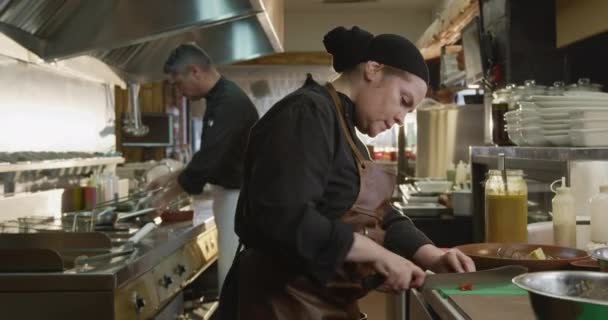 忙しいレストランのキッチンで働く白人女性の料理人の側面図 野菜をスライス バックグラウンドで働く彼女の同僚 — ストック動画