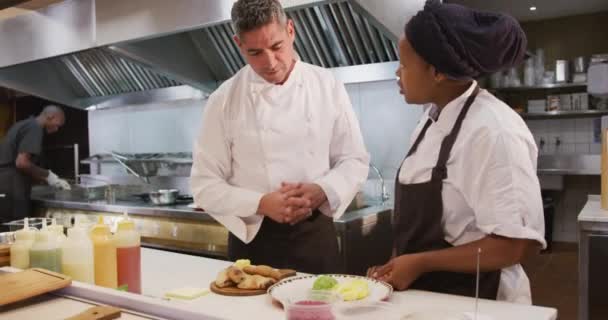 忙しいレストランキッチンで働く白人男性シェフのフロントビュー アフリカ系アメリカ人女性料理人と一緒に提供するプレートをチェック — ストック動画
