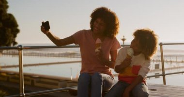 Güneşli bir günde oğluyla birlikte deniz kenarında eğlenen, dondurma yiyen, selfie çeken, yavaş çekim yapan melez bir kadın.