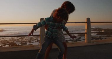 Gün batımında güneşli bir günde oğluyla birlikte deniz kenarında vakit geçiren, kaykay yapan, yavaş çekim yapan melez bir kadının ön görüntüsü.