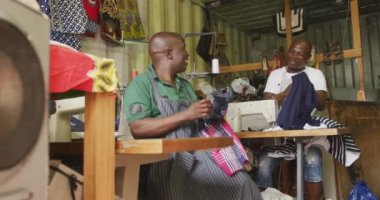 Front view of two African male tailors in a township workshop, sitting by sewing machines, holding fabric and talking, slow motion