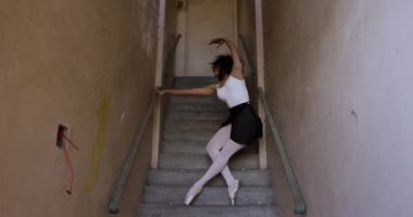 Boş bir depoda dans eden ve merdivenlerde gerinen melez bir kadın balerin düşük açılı görüntüsü.