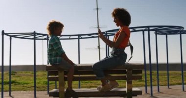 Güneşli bir günde güneşli bir günde oğluyla çocuk parkında eğlenen, bir bankta oturan, bir çocuğun fotoğrafını çeken anne ve oğlunun yan görüntüsü.