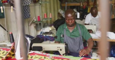 Kasaba atölyesindeki iki Afrikalı erkek terzinin önü. Dikiş makinelerinin yanında oturuyorlar, kumaş tutuyorlar, dikiş dikiyorlar, ağır çekim.