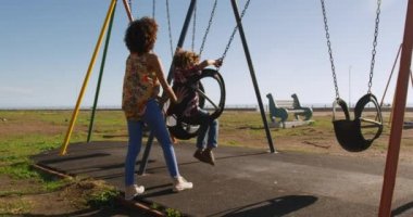 Mavi gökyüzü güneşli bir günde oğluyla çocuk parkında vakit geçiren salıncakta sallanan, ağır çekimde oynayan melez bir kadının yan görüntüsü.