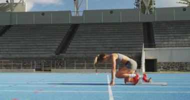 Bir spor stadyumunda antrenman yapan beyaz bir bayan sporcunun yan görüntüsü. Koşu bloklarından, ağır çekimde koşmaya başlıyor.