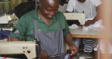 Kasaba atölyesindeki iki Afrikalı erkek terzinin önü, dikiş makinelerinin yanında oturmuş, kumaş tutuyor, kesiyor ve dikiyor, yavaş çekim