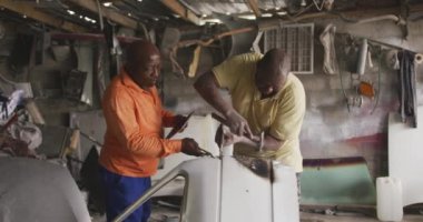 Şehirdeki bir atölye atölyesinde çalışan Afrikalı bir erkek, elinde tablet bir bilgisayar, çalışanının yanında duruyor, talimatlar veriyor, diğeri arabayı tamir etmek için çekiç kullanıyor, yavaş çekim.