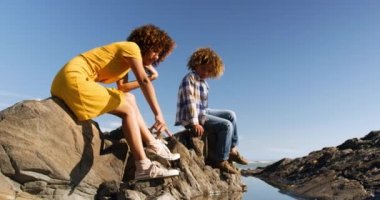 Mavi gökyüzü ile güneşli bir günde oğluyla birlikte deniz kenarında eğlenen, kayalara bakan ve ağır çekim gösteren melez bir kadının yan görüntüsü.