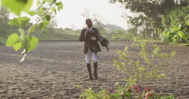 ドレッジホースに乗る前に鞍を保持パドックを歩くアフリカ系アメリカ人の男のスマートな服装のフロントビュー スローモーション — ストック動画