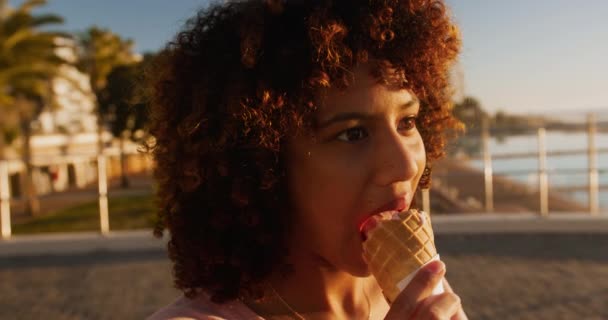 青い空と晴れた日に海辺で時間を楽しむ混合レースの女性の近くにサイドビュー アイスクリームを食べます スローモーション — ストック動画