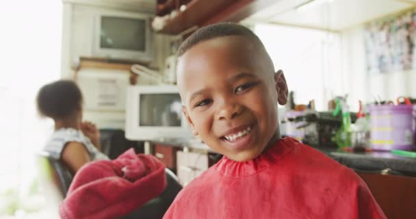髪の毛のサロンで幸せなアフリカの少年の肖像画は 髪の毛の後にカメラを見て 彼のお母さんは背景で待っている 遅い動き — ストック動画