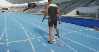Spor stadyumunda antrenman yapan karışık ırktan bir erkek sporcunun arka görüntüsü. Koşu bloklarında yarışmaya hazırlanıyor, ağır çekimde.