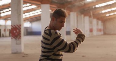 Boş bir depoda dans eden ve kolları havada dönüp duran beyaz erkek bir balerin ön planda.