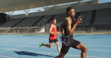 Bir spor stadyumunda antrenman yapan beyaz ve Afrikalı Amerikalı erkek sporcuların yan görüntüsü. Koşu pistinde birbirleriyle yarışıyorlar, ağır çekimde.