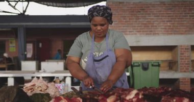 Front view of an African female butcher wearing a headscarf in a township workshop, cutting fresh meat with knife, slow motion
