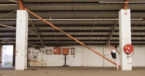 一个混血的女芭蕾舞演员在一个空荡荡的仓库里练习 伸展着身体靠在柱子上 腿靠在横梁上 动作缓慢的侧视图 — 图库视频影像