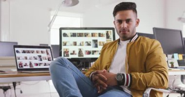 Portrait of a happy mixed race man working in a creative office, sitting by a desk with computers, looking at camera, smiling