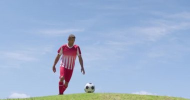 Side view of Caucasian male football player wearing a team strip, training at a sports field in the sun, kicking a football, in slow motion