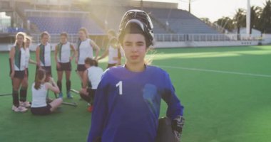 Kafkasyalı genç bir bayan hokey oyuncusunun portresi, kaleci, bir spor stadyumunda başlık ve pedlerle duruyor, kameraya bakıyor ve güneşe gülümsüyor, takım arkadaşları arka planda duruyor, yavaş çekim.