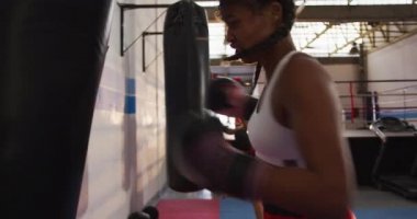 Bir boks salonunda uzun, koyu renk saç eğitimi almış, boks eldiveni giymiş ve kum torbasını yumruklamış, arka planda antrenman yapan bir kadın boksörün yan görüntüsü.