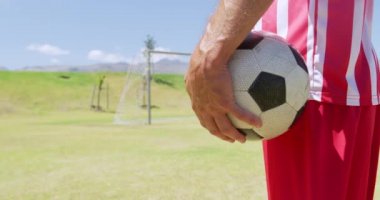 Kafkasyalı bir futbolcunun orta kesiminde takım elbiseli, güneşin altında bir spor sahasında antrenman yapan, mola sırasında ayakta duran ve elinde ağır çekimde futbol topu tutan. Stadyumda Koşu ve Spor Eğitimi.