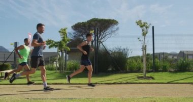 Bir spor sahasında birlikte çim pistinde koşan çok ırklı bir erkek koşucu grubunun yan görüntüsü. Stadyumda ağır çekimde Koşu ve Spor Eğitimi