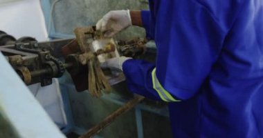 Bir fabrikada çalışan, kriket topları yapan, kriket topunun çekirdeği için doldurulmuş bir kalıbı makinenin etrafında döndüren eldiven ve tulum giyen bir adamın yan görüntüsü.