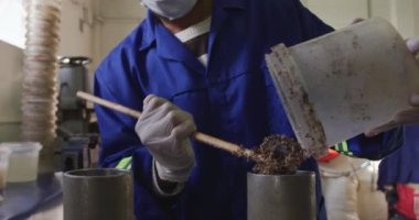 Yüz maskesi takan, eldivenli ve tulumlu melez bir adamın ön tarafı. Bir fabrikada çalışıyor. Kriket topları yapıyor, topların çekirdeği için bir karışım hazırlıyor.