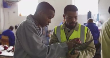 Fabrikada çalışan, kriket topları yapan, fabrika atölyesindeki ürünleri tartışan iki Afrikalı Amerikalının ön görüntüsü. Bir adam yüksek görüş mesafesi olan bir yelek giyiyor ve elinde bir pano var.