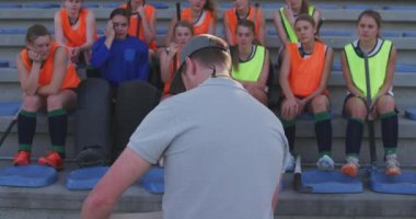 Kafkasyalı bir erkek koçun, genç bir beyaz kadın hokey takımına, stadyum koltuklarında ağır çekimde oturmalarını öğretmesi.